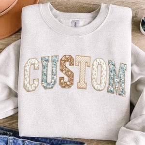Pu&ograve; includere: Una felpa grigio chiaro con la scritta "CUSTOM" applicata sul petto. Le lettere sono in stile collegiale, con dettagli in tessuto floreale e fantasia nei toni del blu, marrone e crema.