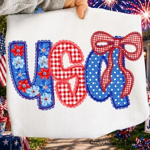 May include: White fabric with the text "USA" in red, white, and blue. The letter "U" is blue with red and white flowers, the "S" is red gingham, and the "A" is blue with white polka dots and a red gingham bow. The design is trimmed with lace.