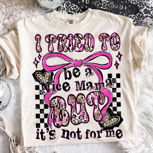 May include: Cream-colored t-shirt with the text "I tried to be a Nice Mama BUT it's not for me" in pink and leopard print. The design includes a pink bow, stars, and a checkered pattern.