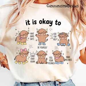 May include: Cream-colored t-shirt featuring a graphic with cartoon Highland cows and the text "it is okay to." The cows are depicted in various poses with phrases like "Be Yourself" and "Make Mistakes."