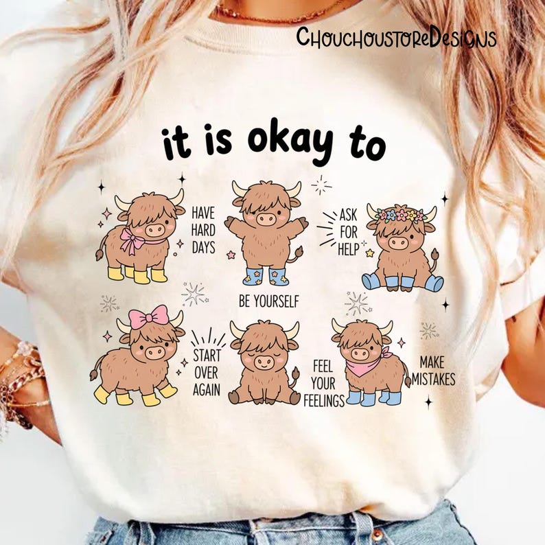 May include: Cream-colored t-shirt featuring a graphic with cartoon Highland cows and the text "it is okay to." The cows are depicted in various poses with phrases like "Be Yourself" and "Make Mistakes."