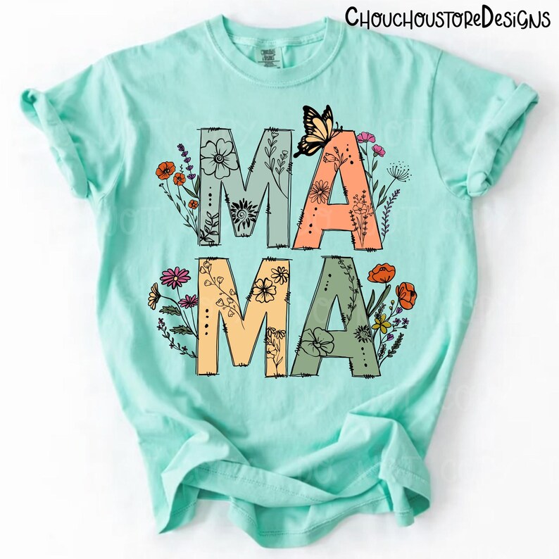 May include: A light teal t-shirt with the word "MAMA" in large, colorful letters. The letters are decorated with floral designs and a butterfly. The top is a comfortable, casual style.