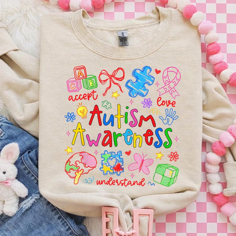 May include: A beige sweatshirt with a colorful graphic promoting Autism Awareness. The design includes puzzle pieces, a ribbon, and the words "accept," "love," and "understand." The phrase "Autism Awareness" is prominently displayed in various colors.