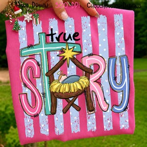 May include: A bright pink t-shirt featuring a graphic with the text true STORY. The design includes a teal cross, a yellow star, and a manger scene with a baby in a yellow crib. The background has light blue vertical stripes with white dots.