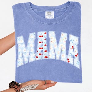 May include: A light blue Comfort Colors t-shirt with the word "MAMA" in a floral and cherry design. The letters are outlined with a white lace trim. The shirt is held up by a person.