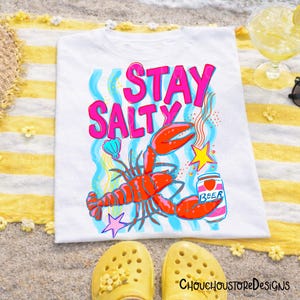 Puede incluir: Camiseta blanca con un gr&aacute;fico colorido que dice "Stay Salty". El dise&ntilde;o incluye una langosta roja, una lata de cerveza y una estrella. La camiseta est&aacute; sobre una toalla a rayas amarillas y blancas, con zapatos amarillos en primer plano.