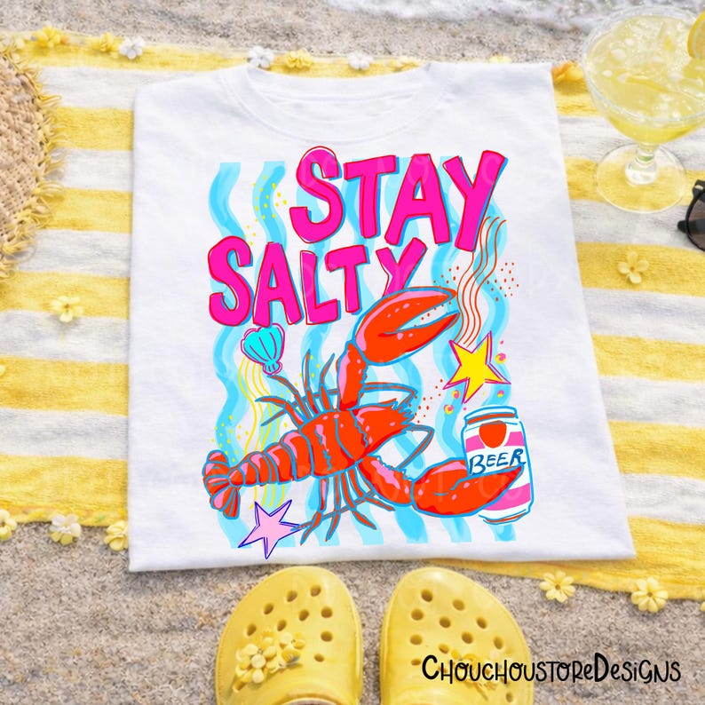 Puede incluir: Camiseta blanca con un gr&aacute;fico colorido que dice "Stay Salty". El dise&ntilde;o incluye una langosta roja, una lata de cerveza y una estrella. La camiseta est&aacute; sobre una toalla a rayas amarillas y blancas, con zapatos amarillos en primer plano.