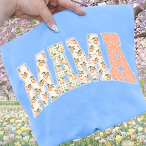 May include: A light blue sweatshirt with the word "MAMA" appliqued across the front. The letters are in a patchwork style, with peach and floral patterns. The letter "A" is a solid peach color with white polka dots.