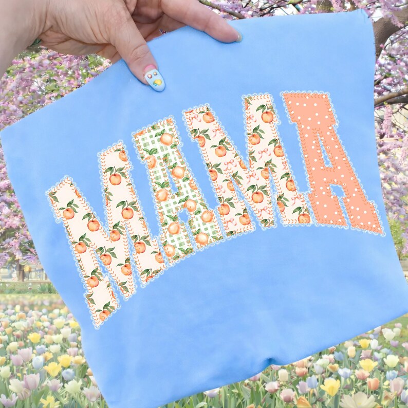 May include: A light blue sweatshirt with the word "MAMA" appliqued across the front. The letters are in a patchwork style, with peach and floral patterns. The letter "A" is a solid peach color with white polka dots.