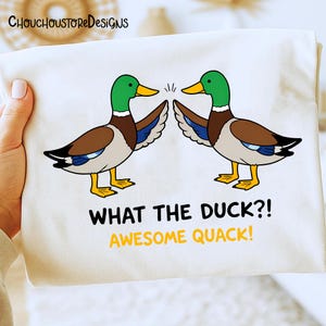 May include: A white t-shirt featuring a cartoon design of two ducks with green heads, brown bodies, and yellow beaks and feet. The text "WHAT THE DUCK?! AWESOME QUACK!" is printed below the ducks.