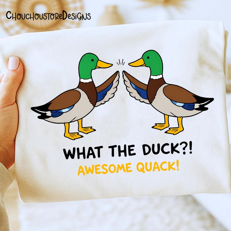 May include: A white t-shirt featuring a cartoon design of two ducks with green heads, brown bodies, and yellow beaks and feet. The text "WHAT THE DUCK?! AWESOME QUACK!" is printed below the ducks.