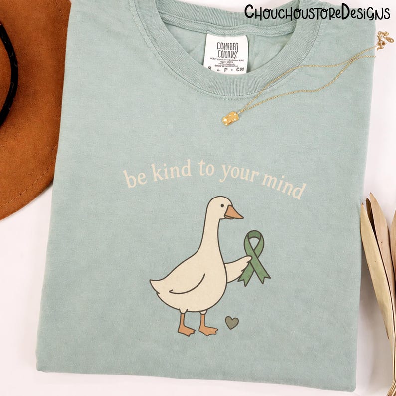 May include: A light blue t-shirt with the phrase "be kind to your mind" printed above a cartoon goose holding a green ribbon. A small heart is next to the goose. A gold necklace is draped across the shirt.