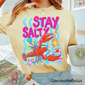 Puede incluir: Camiseta amarilla clara con un dise&ntilde;o gr&aacute;fico colorido. Presenta una langosta roja, una lata de cerveza y la frase "STAY SALTY" en rosa. L&iacute;neas onduladas azules, una concha y una estrella completan el dise&ntilde;o. Prenda informal con tem&aacute;tica playera.