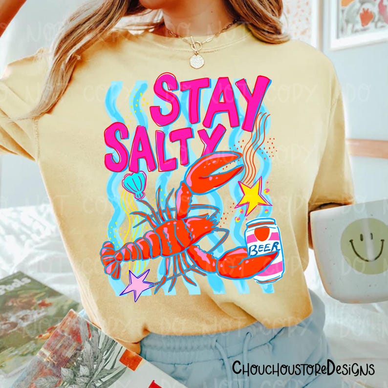Puede incluir: Camiseta amarilla clara con un dise&ntilde;o gr&aacute;fico colorido. Presenta una langosta roja, una lata de cerveza y la frase "STAY SALTY" en rosa. L&iacute;neas onduladas azules, una concha y una estrella completan el dise&ntilde;o. Prenda informal con tem&aacute;tica playera.