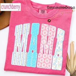 May include: A pink t-shirt with the word "MAMA" in a decorative font. The letters are filled with various patterns, including blue polka dots, floral designs, and striped patterns. The shirt has the "Comfort Colors" brand tag.