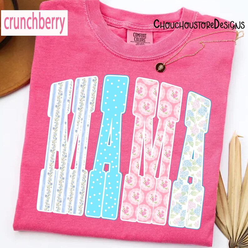 May include: A pink t-shirt with the word "MAMA" in a decorative font. The letters are filled with various patterns, including blue polka dots, floral designs, and striped patterns. The shirt has the "Comfort Colors" brand tag.