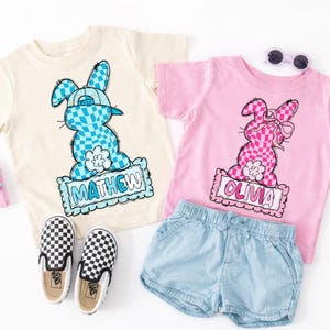 May include: Two children's t-shirts, one cream and one pink, each with a checkered bunny graphic and a name. The cream shirt has a blue bunny with the name "MATHEW". The pink shirt has a pink bunny with the name "OLIVIA". Also pictured are denim shorts and checkered shoes.