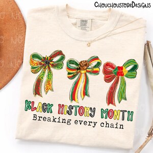 Op de afbeelding: Cr&egrave;mekleurig T-shirt met drie decoratieve strikken in rood, groen en geel. Het shirt heeft de tekst "Black History Month" en "Breaking every chain" in kleurrijke letters. Een ketting en een hoed zijn zichtbaar op de achtergrond.