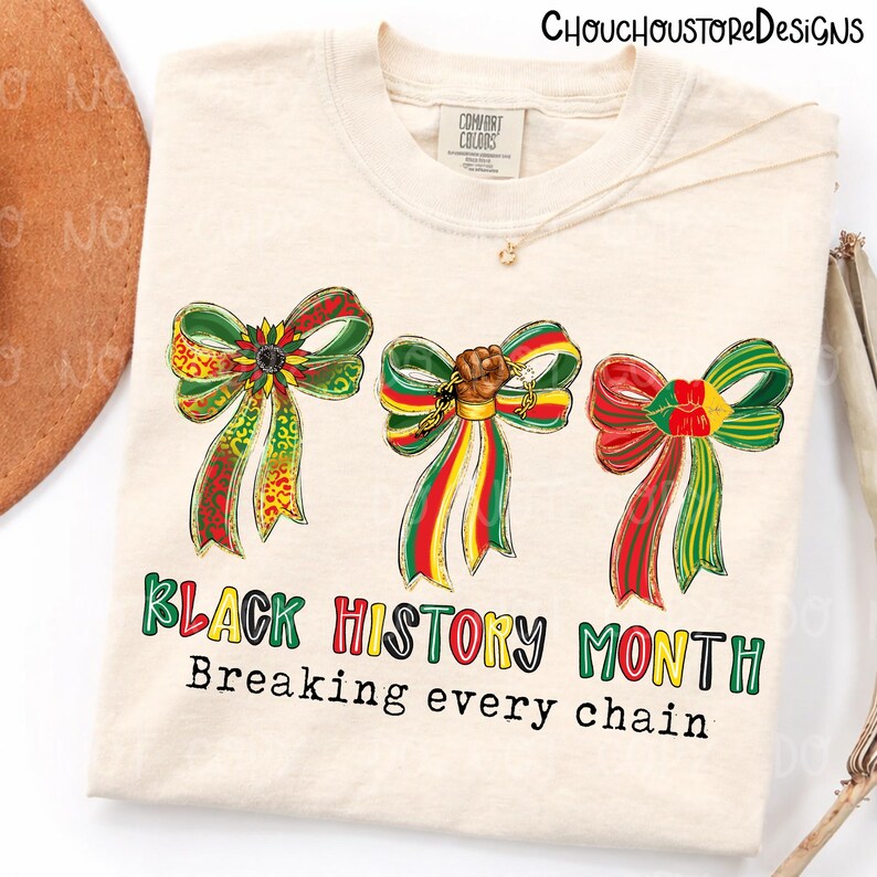 Op de afbeelding: Cr&egrave;mekleurig T-shirt met drie decoratieve strikken in rood, groen en geel. Het shirt heeft de tekst "Black History Month" en "Breaking every chain" in kleurrijke letters. Een ketting en een hoed zijn zichtbaar op de achtergrond.