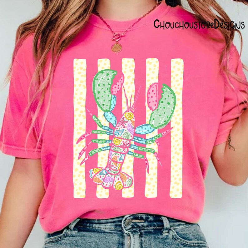 May include: Pink t-shirt featuring a colorful lobster design with a striped background. The lobster has green and pink claws, and its body is decorated with various patterns. The text "ChouchouStoreDesigns" is visible in the upper right corner.