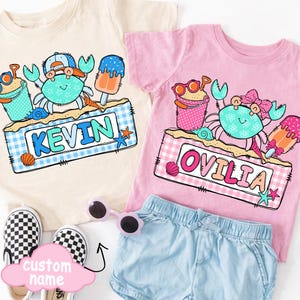 May include: Two children's t-shirts, one cream and one pink, featuring crab designs with sunglasses, ice cream, and the names "KEVIN" and "OVILIA". The shirts are paired with denim shorts and checkered slip-on shoes. A pink cloud-shaped sign says "custom name".