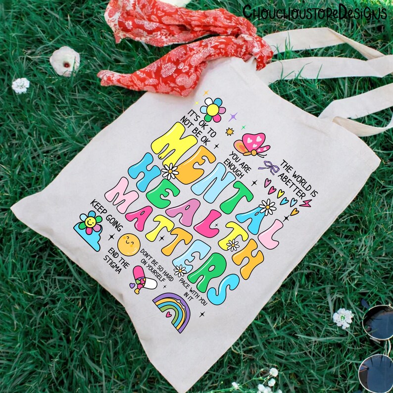 May include: A canvas tote bag with the words "MENTAL HEALTH MATTERS" in colorful, bubble letters. The bag also features various graphics and phrases like "It's ok to not be ok" and "You are enough". A red scarf and sunglasses are in the background.