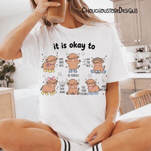 May include: White t-shirt with the text "it is okay to" and illustrations of cartoon Highland cows. Each cow has a different accessory and a phrase like "Have Hard Days" or "Be Yourself".