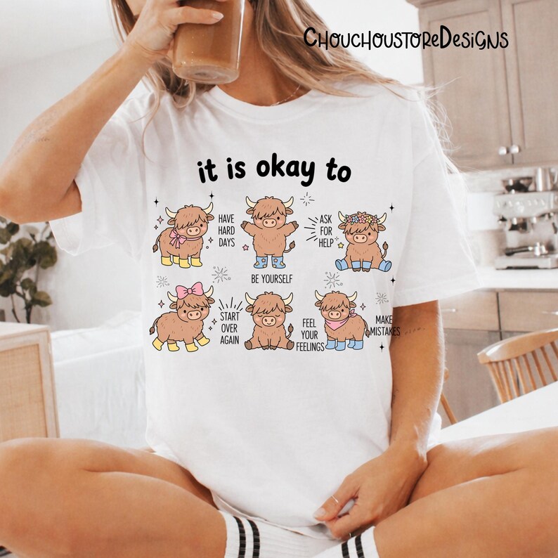 May include: White t-shirt with the text "it is okay to" and illustrations of cartoon Highland cows. Each cow has a different accessory and a phrase like "Have Hard Days" or "Be Yourself".