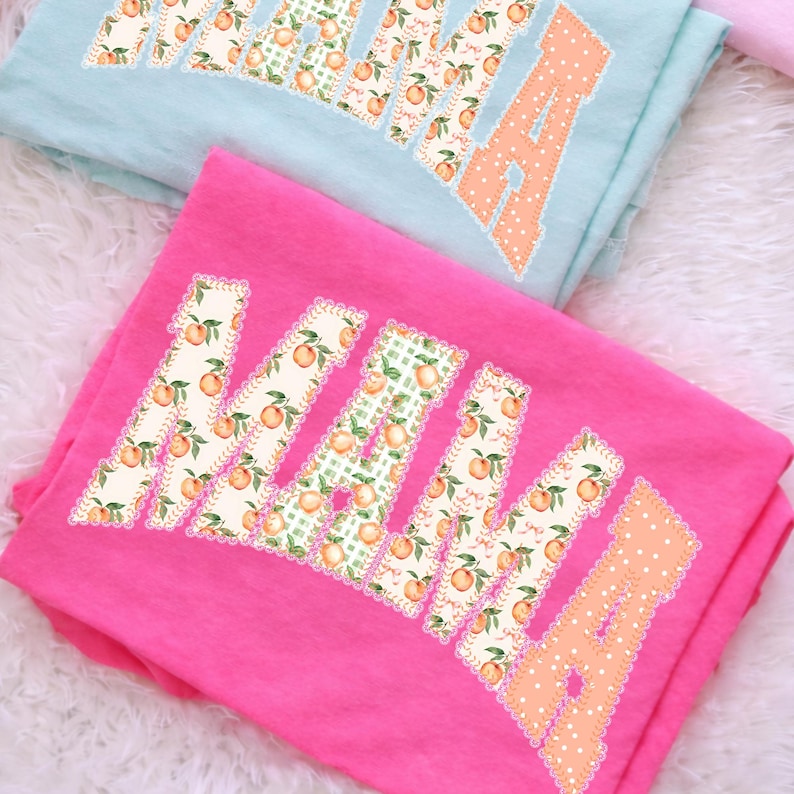 May include: Two t-shirts, one pink and one light blue, are folded. Each shirt features the word "MAMA" in large, decorative letters. The letters are filled with a peach and leaf pattern, with a scalloped edge.