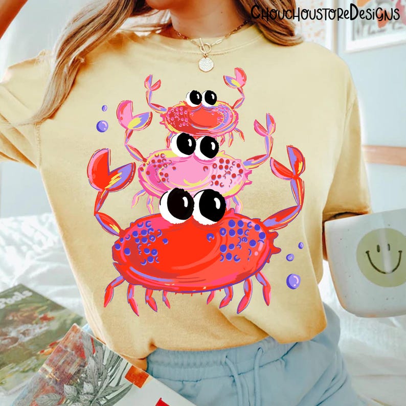 May include: A light yellow sweatshirt featuring a stack of three cartoon crabs in red, pink, and purple. The crabs have large black eyes and are surrounded by purple bubbles. The sweatshirt has the text "ChouChouStoreDesigns" at the top.