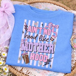 May include: A light blue sweatshirt with the text "Ain't No Hood Like Motherhood" in pink and purple. The design includes illustrations of coffee cups, bows, flowers, and lightning bolts. The shirt is folded in a wicker basket.