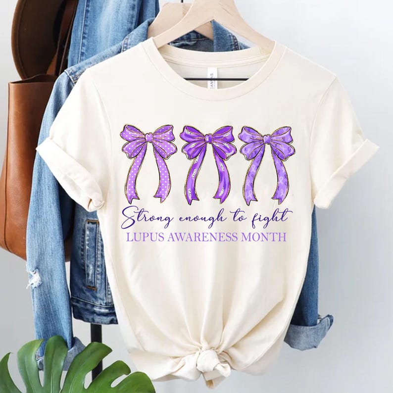 K&ouml;nnte beinhalten: Cremefarbenes T-Shirt mit drei lila Schleifen mit wei&szlig;en Punkten und goldenen Akzenten. Der Text "Strong enough to fight" und "Lupus Awareness Month" ist unter den Schleifen aufgedruckt.