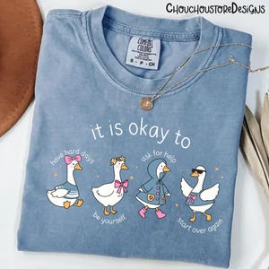 May include: A light blue Comfort Colors t-shirt with the text "it is okay to" and four cartoon geese. Each goose has a different accessory and phrase: "have hard days", "be yourself", "ask for help", and "start over again". A gold necklace is draped over the shirt.