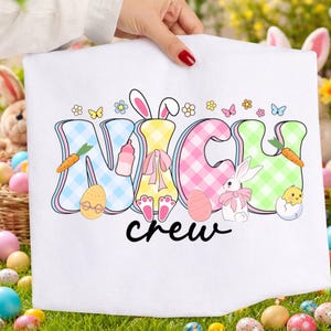 May include: White Easter-themed towel with the words "NICH CREW" in colorful, checkered letters. The design includes Easter eggs, bunnies, carrots, and flowers. The towel is held up by a person with red nail polish.