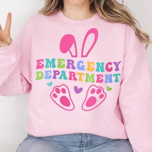 May include: A light pink sweatshirt with the words "EMERGENCY DEPARTMENT" in colorful, retro-style lettering. The design includes pink bunny ears, paw prints, and hearts, perfect for Easter or a medical professional.