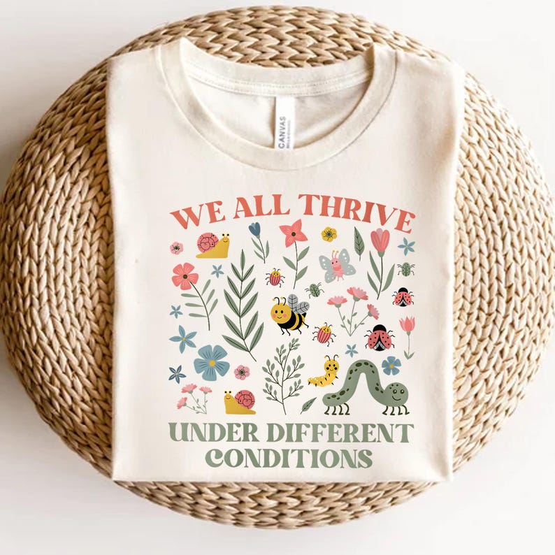 Peut inclure: T-shirt cr&egrave;me avec le texte "WE ALL THRIVE UNDER DIFFERENT CONDITIONS" en rouge et vert. Le t-shirt est orn&eacute; d'illustrations color&eacute;es de fleurs, d'insectes et d'un escargot.