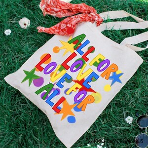 May include: A white canvas tote bag with the words "ALL LOVE FOR ALL" in colorful, block letters and star shapes. The bag has long, natural-colored handles and is lying on green grass. A red and white patterned scarf is draped nearby.