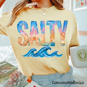 May include: Yellow t-shirt with the word "SALTY" in colorful, textured lettering above blue ocean waves. The shirt has a relaxed fit and is likely made of soft material. The design evokes a beach or coastal theme.