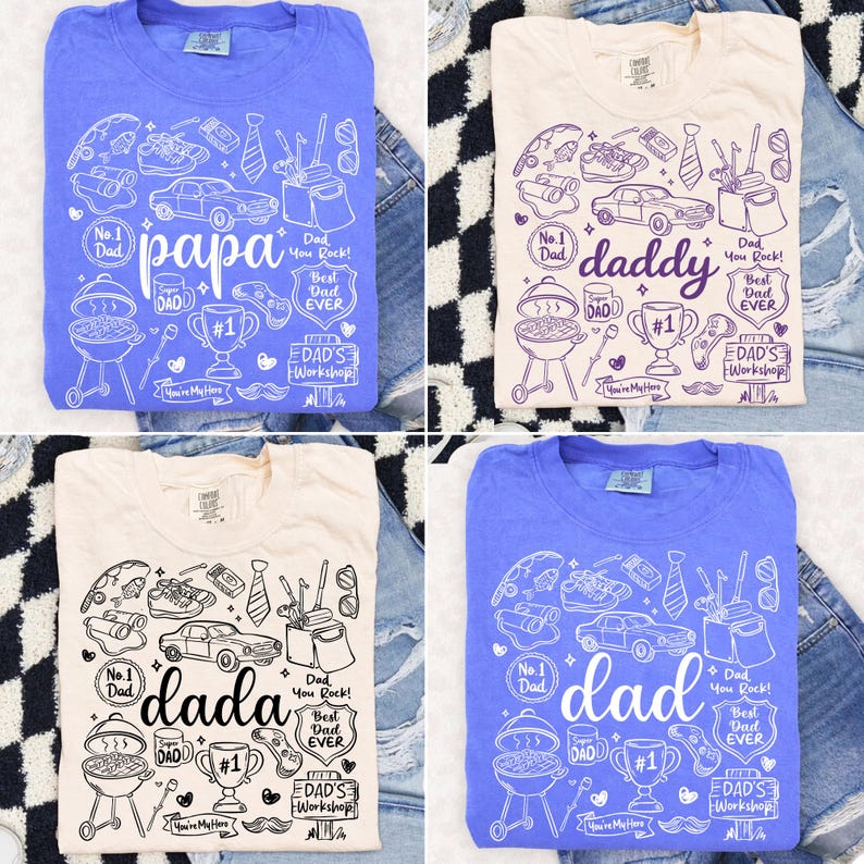 May include: Four t-shirts in blue and cream colors, each with a unique graphic design. The designs include the words "papa", "daddy", "dada", and "dad", along with various illustrations like a grill, car, and trophy. The shirts are laid out on denim.