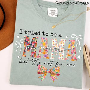 May include: A light green t-shirt with the text "I tried to be a nicer MAMA but It's not for me." The word "MAMA" is decorated with colorful floral patterns. The shirt has a bow graphic at the bottom.
