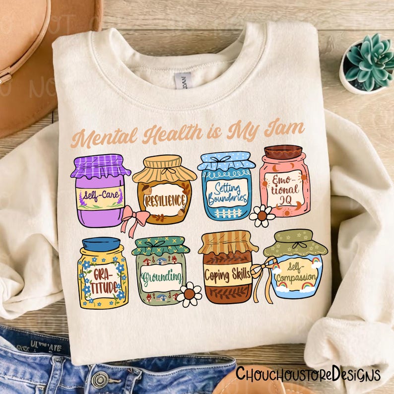 May include: Cream-colored sweatshirt with the phrase "Mental Health is My Jam" and illustrations of jam jars labeled with mental health concepts like "Self-Care" and "Resilience".