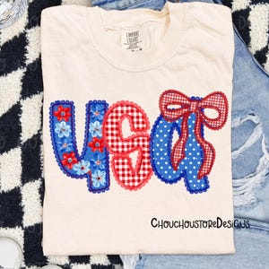May include: Cream-colored t-shirt with the word "USA" in red, white, and blue appliqued letters. The "U" is blue with floral accents, the "S" is red gingham, and the "A" is blue with white polka dots and a red gingham bow.