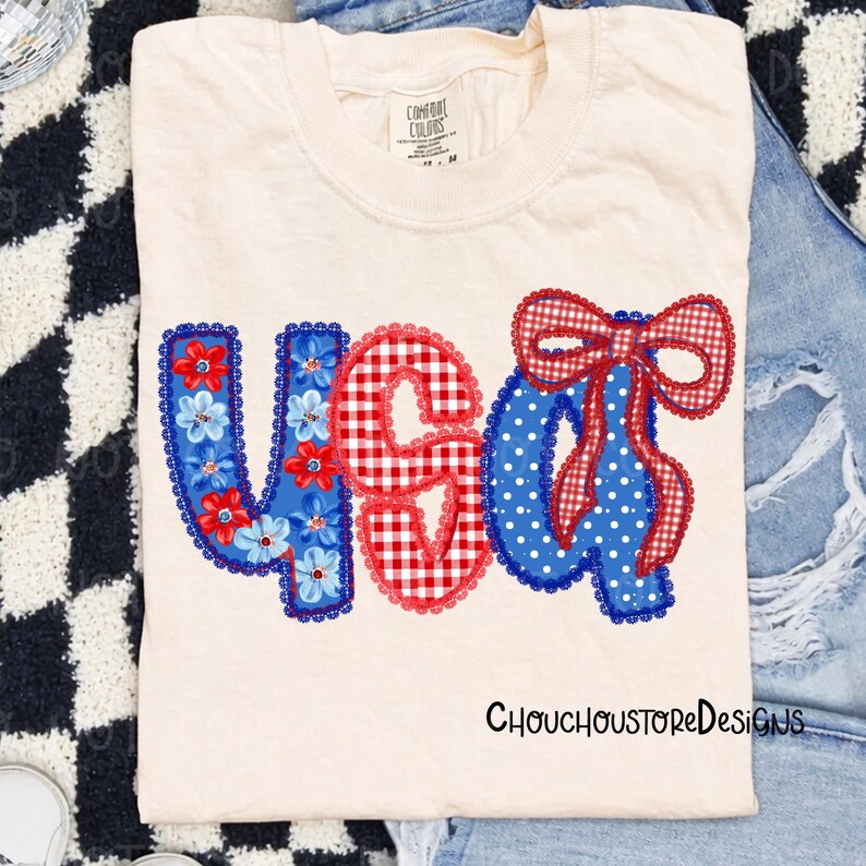 May include: Cream-colored t-shirt with the word "USA" in red, white, and blue appliqued letters. The "U" is blue with floral accents, the "S" is red gingham, and the "A" is blue with white polka dots and a red gingham bow.