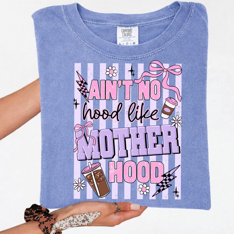 May include: A periwinkle blue t-shirt with the text "Ain't No Hood Like Motherhood" in pink and purple. The design includes lightning bolts, bows, flowers, and coffee cups. The shirt is held up by a person.