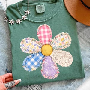 May include: A green t-shirt with a large, colorful flower applique. The flower petals feature various patterns, including gingham, floral prints, and lace. A daisy chain necklace and a brown hat are visible in the background.