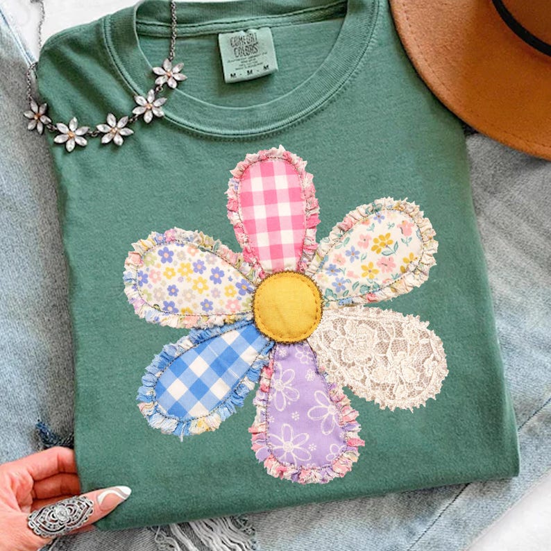 May include: A green t-shirt with a large, colorful flower applique. The flower petals feature various patterns, including gingham, floral prints, and lace. A daisy chain necklace and a brown hat are visible in the background.