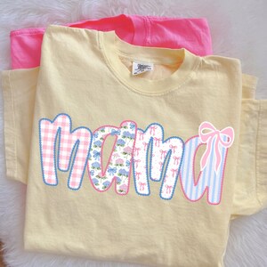 May include: A light yellow t-shirt with the word "mama" in a patchwork design. The letters feature gingham, floral, and striped patterns in pink and blue, outlined in blue. A pink t-shirt is visible in the background.