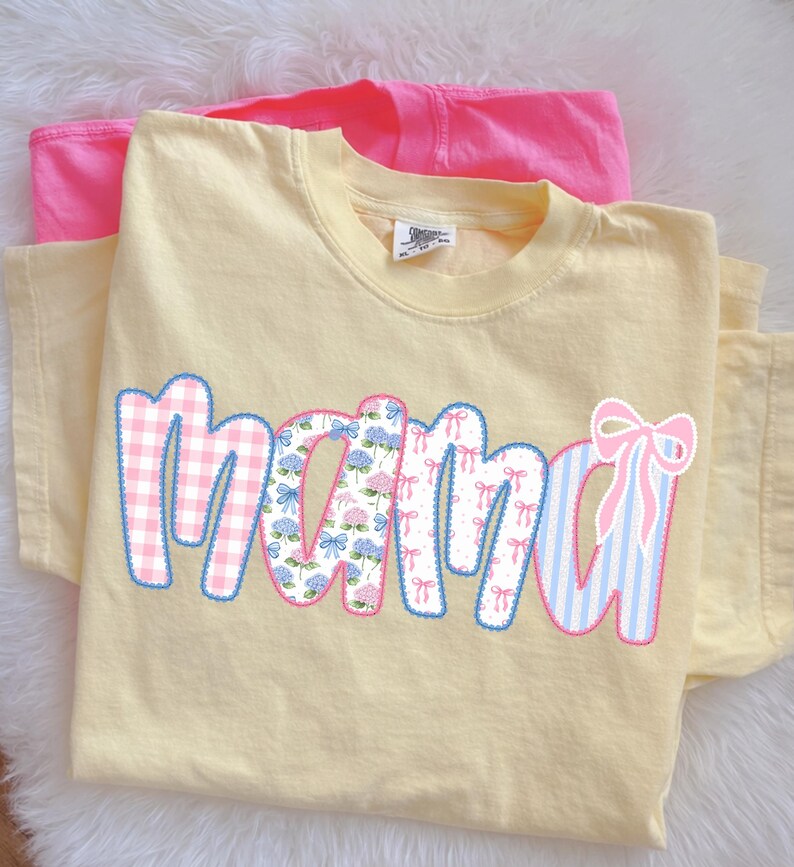 May include: A light yellow t-shirt with the word "mama" in a patchwork design. The letters feature gingham, floral, and striped patterns in pink and blue, outlined in blue. A pink t-shirt is visible in the background.