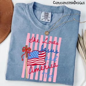 May include: A light blue Comfort Colors t-shirt with the text "She Loves Jesus and America Too" in red and blue script. The design includes a cross, American flag, and pink vertical stripes.