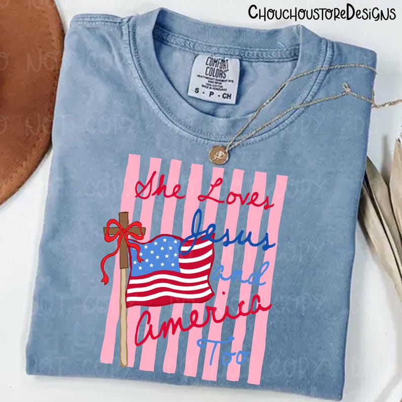 May include: A light blue Comfort Colors t-shirt with the text "She Loves Jesus and America Too" in red and blue script. The design includes a cross, American flag, and pink vertical stripes.